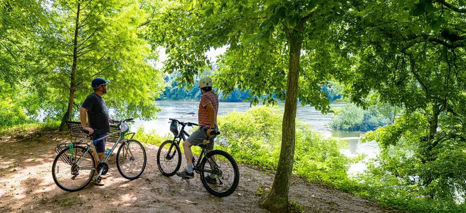 Two bicyclists resting under the shade next to a river.